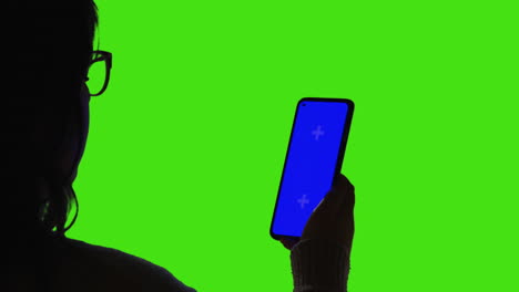 close up over the shoulder silhouette shot of woman wearing glasses looking at blue screen mobile phone against green screen background