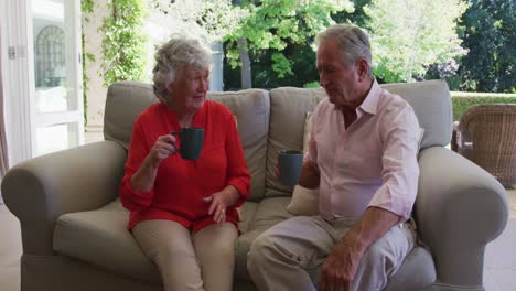 happy caucasian senior couple on couch in sunny living room talking and drinking cups of coffee