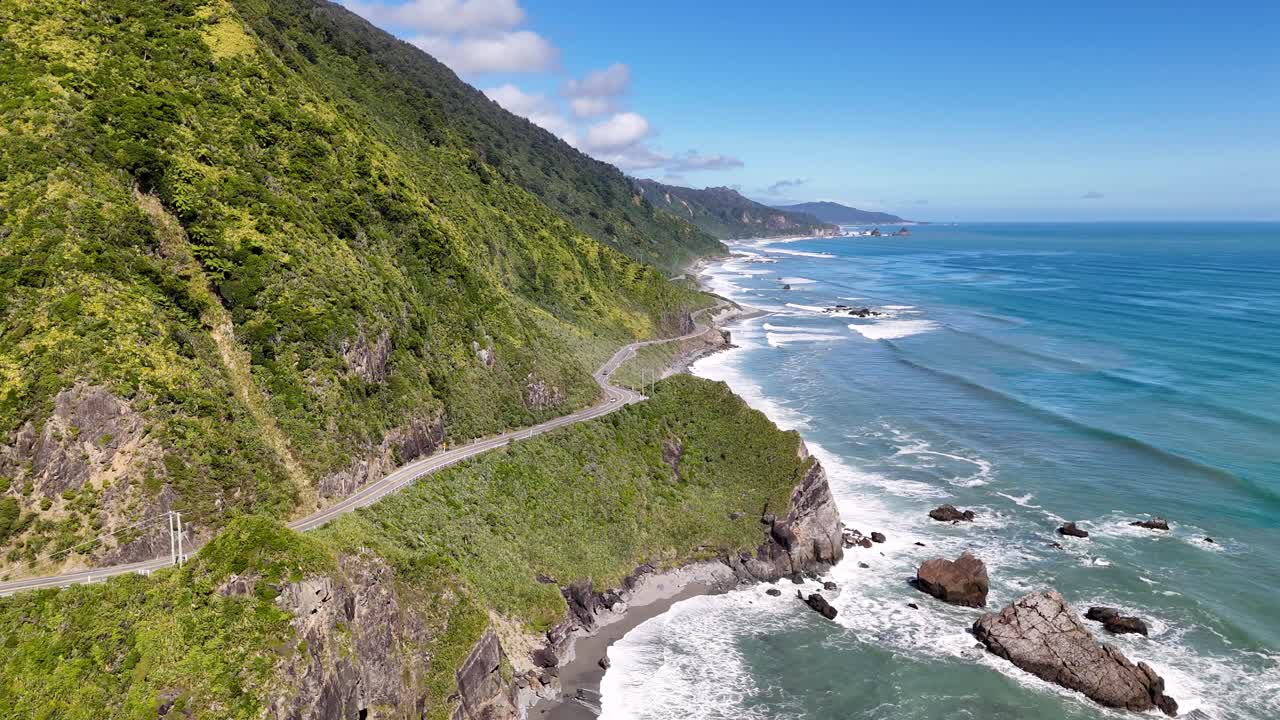 A 4K drone pans around a cliff to reveal a winding coastal road in New Zealand’s South Island. Mountains, ocean, and rugged cliffs create a dramatic aerial view