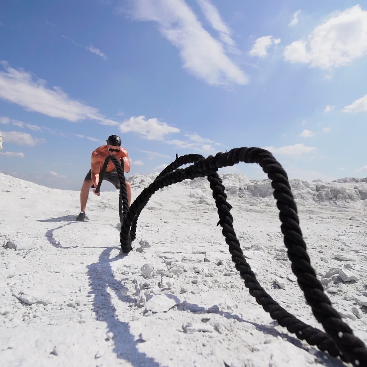 Muscular man with black ropes on white hill under blue sky. Athletic male performs Battle Ropes exercise during an outdoor cross fitness workout. Slow motion.