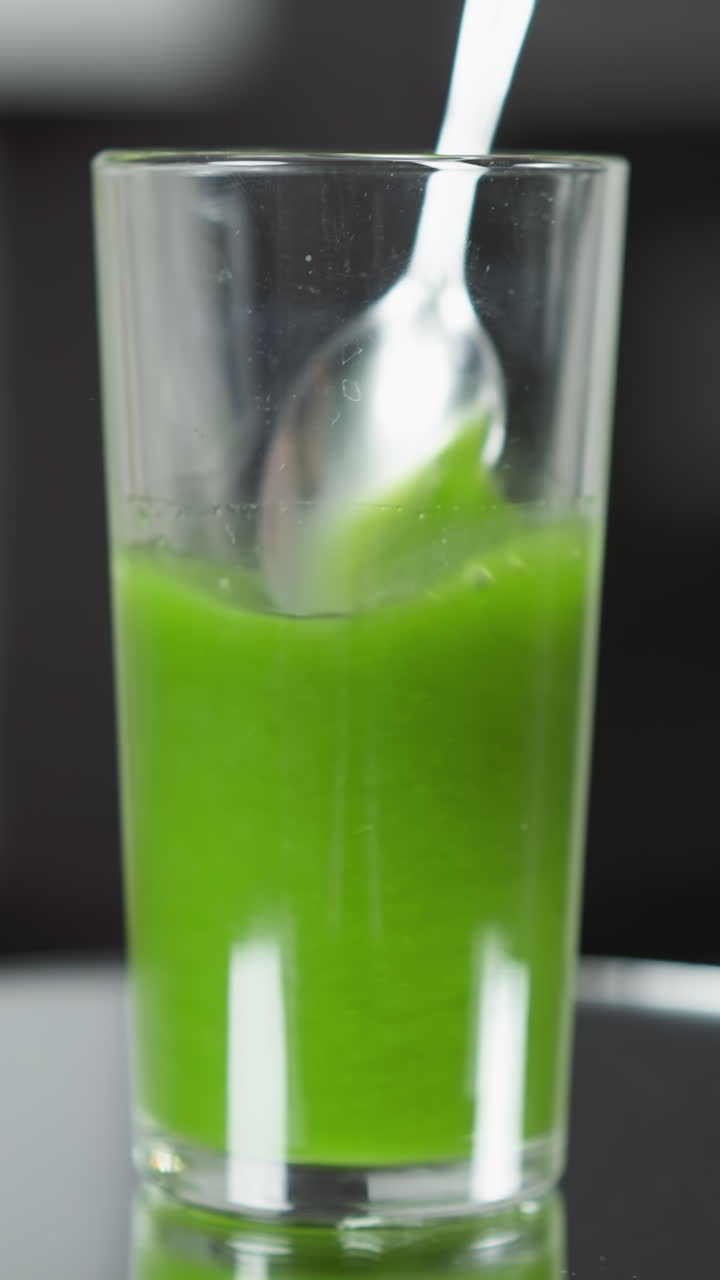 una cuchara intrincadamente diseñada se sumerge en un jugo verde suave y vibrante dentro de un vidrio alto y delgado, un brillo acentuado de luz natural difusa que crea reflejos sutiles en una superficie brillante.