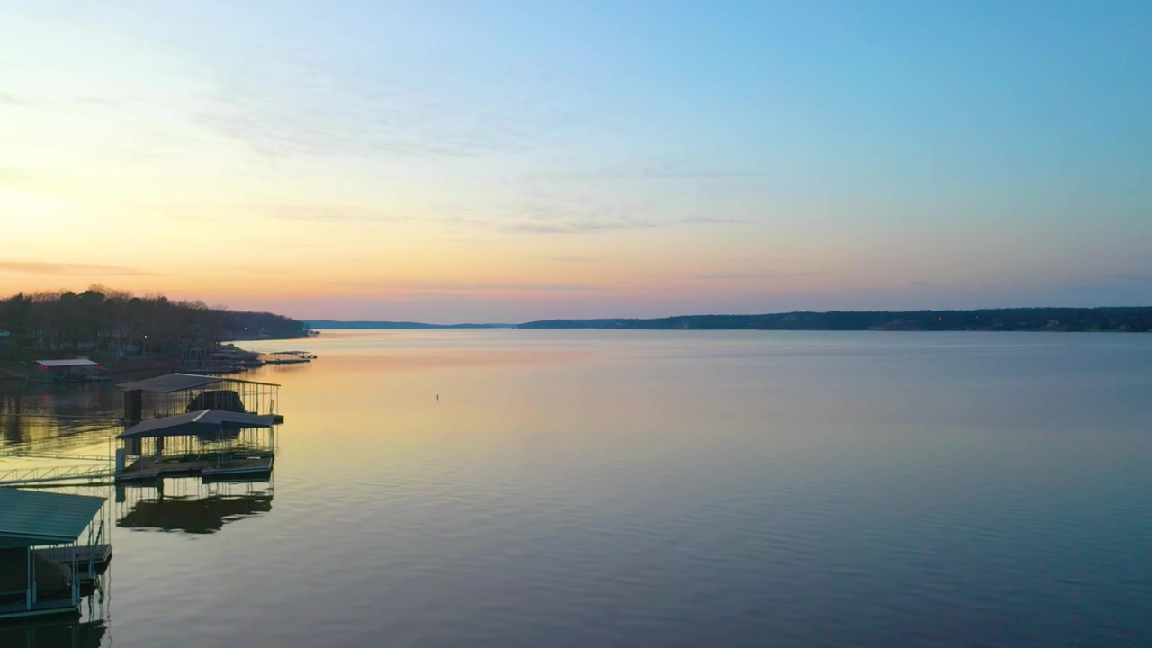 Vista aerea con drone sopra il Lago Beaver al crepuscolo, che mostra l'acqua calma, la costa boscosa dell'Arkansas, le rimesse per barche ormeggiate, le case sul lago e i tenui colori del tramonto sul bacino idrico.