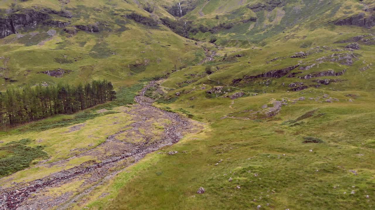 Wide aerial shot, slowly tracking forwards along a hiking trail and a rocky mountain stream on the lush, green valley hillside of Glen Coe, Scotland, United Kingdom, Europe