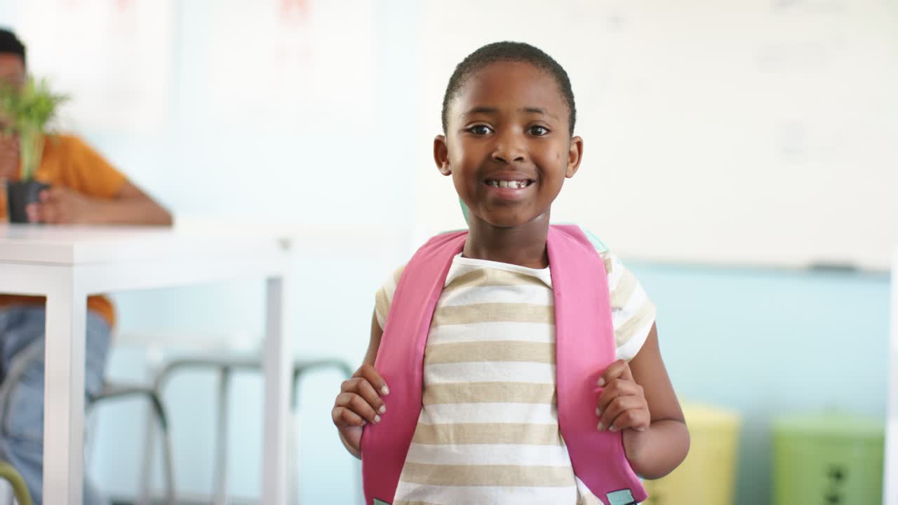 Bambina africana in piedi davanti alla classe, che tiene gli spallacci di uno zaino rosa, sorride e reagisce a un segnale.