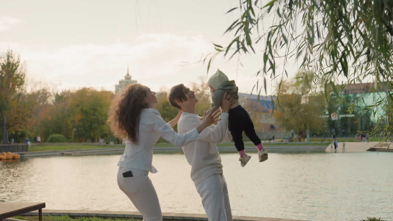 Родители подбрасывают ребенка у воды. Игровая семейная прогулка на выходных с романтической атмосферой. Смеющаяся пара на фоне городского горизонта, скамейка и прибрежные деревья. Непринужденный жизненный момент.