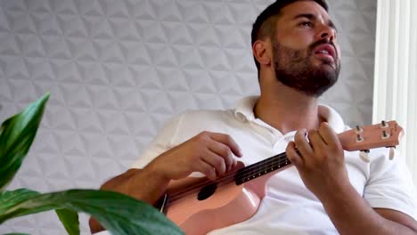 Un-Hombre-De-30-Años-Y-Con-Barba-Está-Tocando-Y-Cantando-Con-Su-Ukelele-Rosa-Junto-A-Una-Gran-Ventana-Y-Cerca-De-Una-Planta-Verde