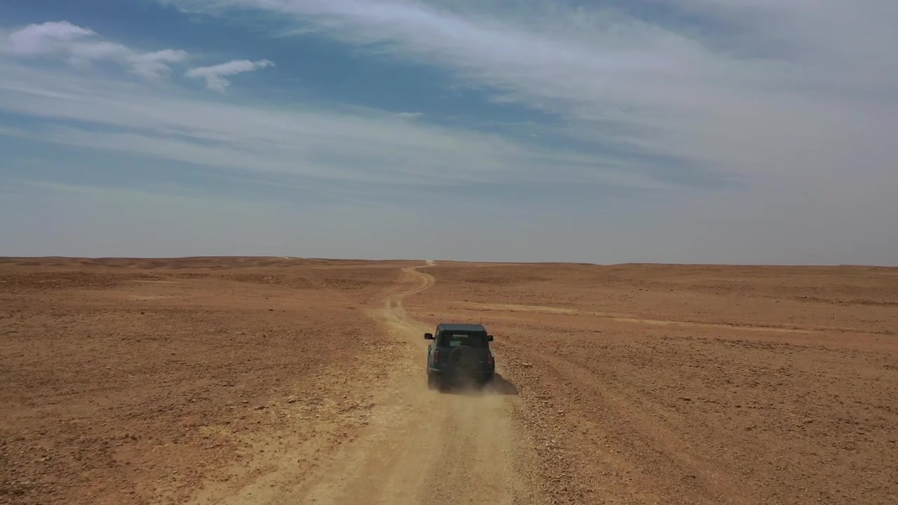 Toma aérea con dron de un coche conduciendo por un camino de tierra aislado a través de la extensa y accidentada meseta desértica cerca del Borde del Mundo, Arabia Saudita, mostrando viajes remotos y aventura