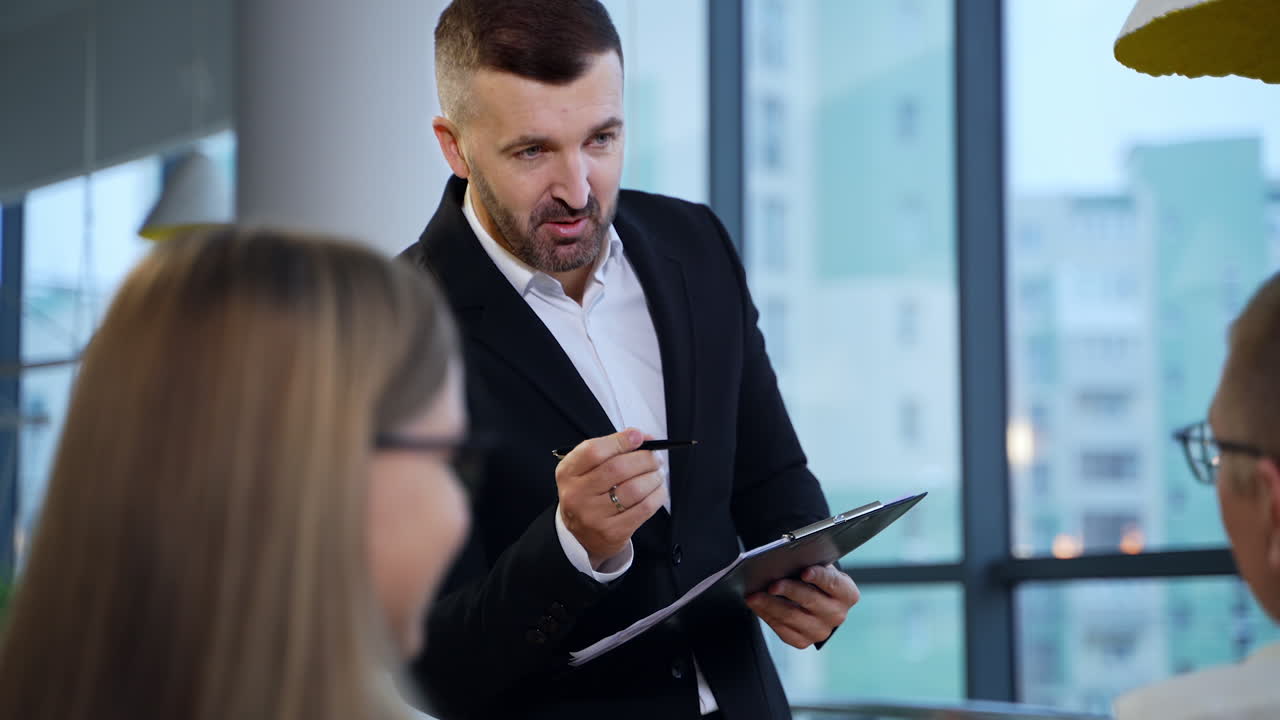 Man in suit standing in front of his colleagues holding a folder and a pen. Smiling man addressing people and asking their opinion. Blurred background.