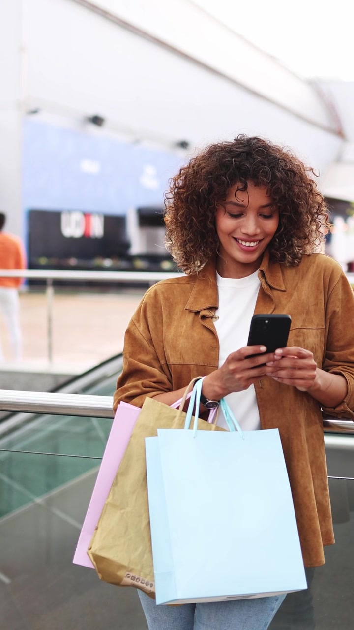 Giovane donna felice con borse della spesa che usa lo smartphone nel centro commerciale