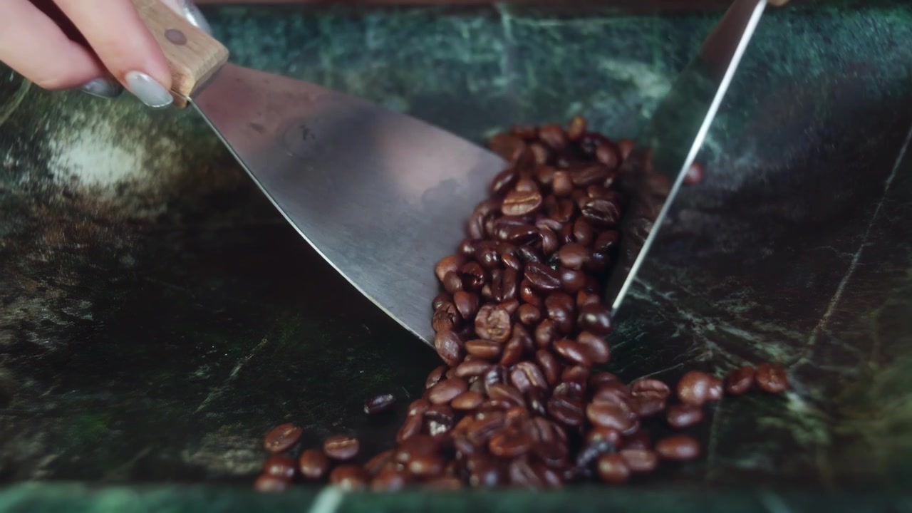 Primo piano di chicchi di caffè tostati a fuoco e liquore alle erbe su una superficie di pietra. Il barista usa raschietti metallici per mescolare la miscela artigianale all'interno di una bacinella di marmo verde