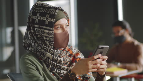 Mujer-De-Negocios-En-Hijab-Y-Máscara-Usando-Teléfono-Inteligente-En-La-Oficina