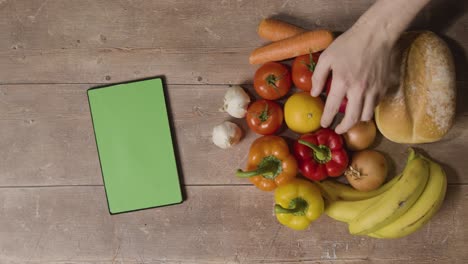 overhead studio shot of hand picking up basic fresh food items with green screen digital tablet on wooden surface 2