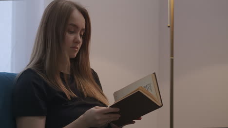 young woman with book in living room at evening closing and turning off floor lamp female portrait in home at weekend resting and self-isolation during lockdown