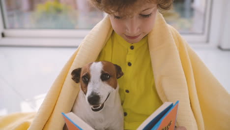 Blonder-Junge-Mit-Lockigem-Haar-Sitzt-Auf-Dem-Boden-Mit-Einer-Decke-Bedeckt-Neben-Seinem-Hund-Beim-Lesen