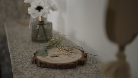 wedding rings displayed on a wooden slab with greenery, next to a vase with a white flower, creating a natural and elegant scene