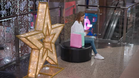 joven sentada en un banco redondo en un centro comercial usando su teléfono, está rodeada de bolsas de compras coloridas con luces festivas y una decoración de estrellas brillantes en el fondo