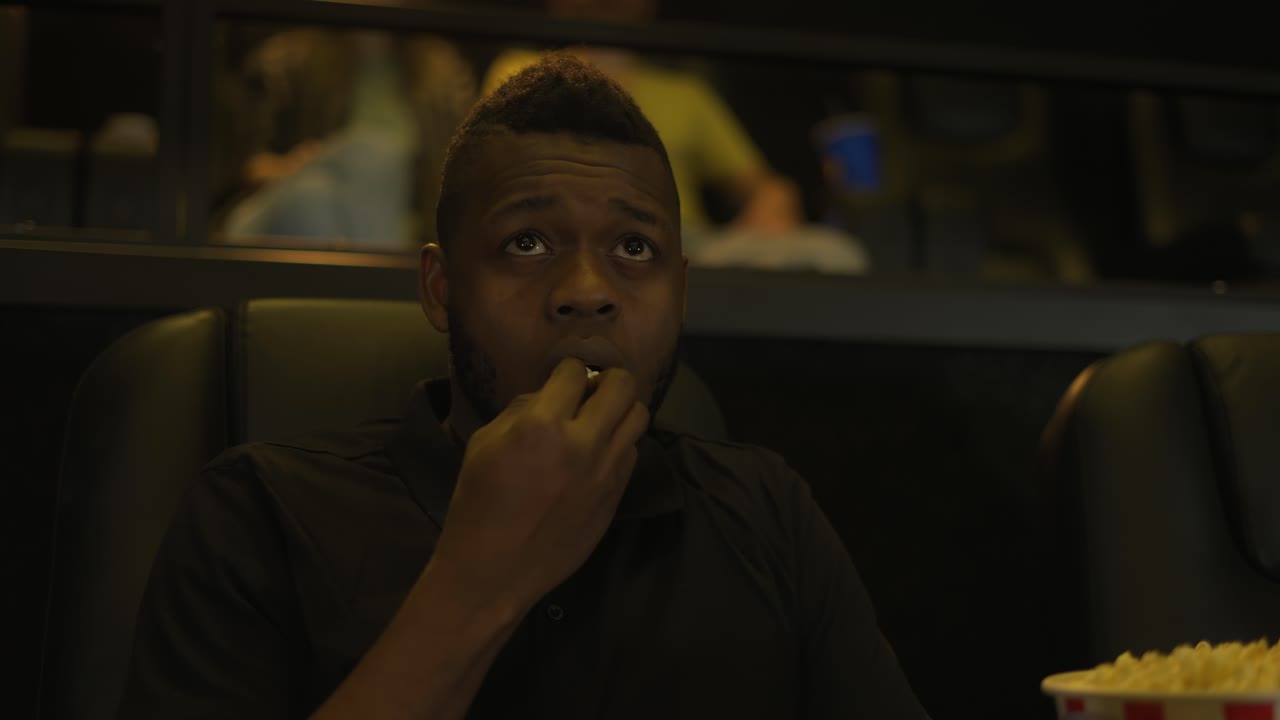 hombre afroamericano sentado en un sillón viendo una película de terror en el cine, cara de miedo