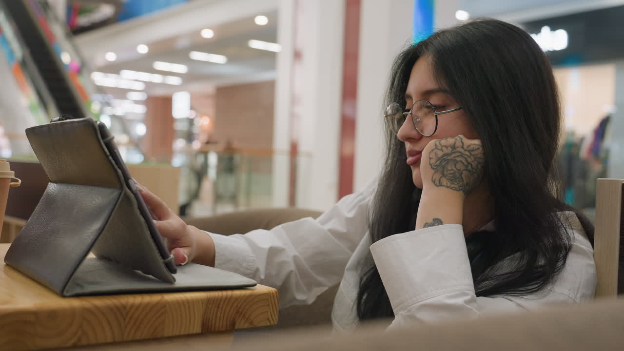 Side view of lady with floral hand tattoo resting head on hand while working on tablet at wooden table in mall, with blurred view of people walking and modern interior in soft lighting background