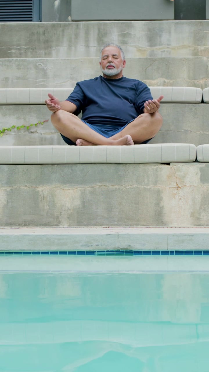 Video verticale: La telecamera rivela un uomo che medita a gambe incrociate su un cuscino da piscina, con una maglietta blu scuro, per rilassarsi.