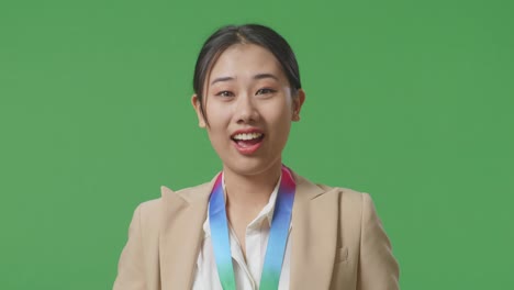 금메달을 입고 카메라를 향해 미소 짓는 아시안 사업가 여성은 스튜디오의 녹색 스크린 배경에서 첫 번째 우승자로 자랑스럽게 승리합니다.