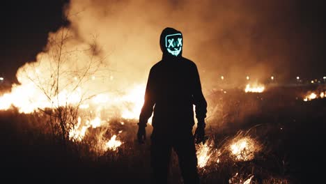 person in glowing mask standing in a field on fire at night