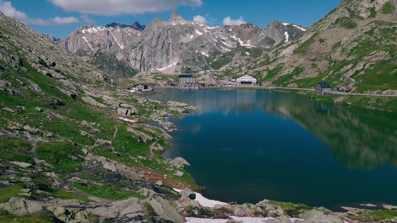 Vista aerea che si avvicina a un lago alpino e a un passo di montagna con edifici storici nelle vicinanze.