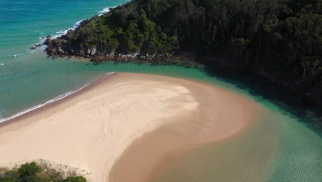 Cinematic-drone-shot-of-the-South-Pacific-Ocean-and-Korogoro-Creek-with-wind-blowing-sand-across-a-sand-bar-at-Hat-Head-New-South-Wales,-Australia