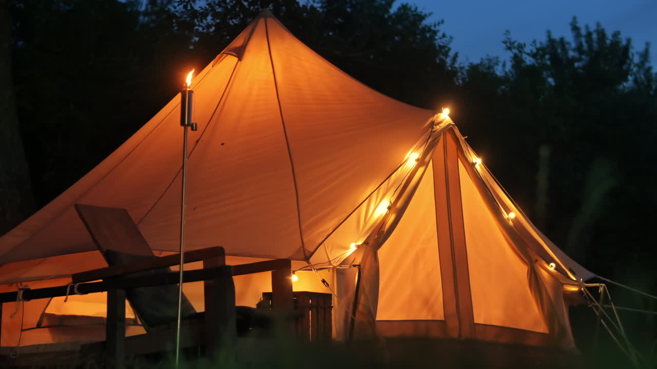 Telt med brændende fakler, lamper og træstole ved glamping, skov rundt om, nat