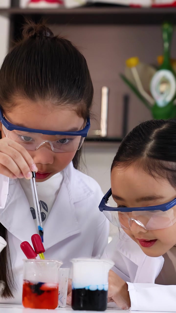 Kaksi aasialaista tyttöä laboratoriotakeissa käyttää pipettejä värikkäiden nesteiden kanssa tieteellisessä kokeessa.