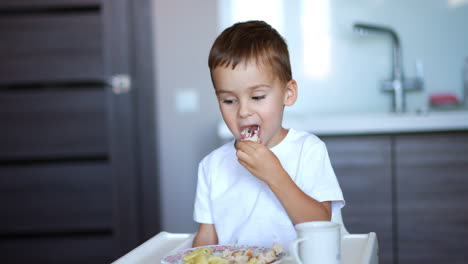 beautiful caucasian baby eating meatballs for lunch. cute toddler in white t-shirt eats at high chair in the kitchen. blurred backdrop.