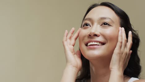 어두운 머리카락을 가진 행복한 아시아 여성이 복사 공간, 슬로우 모션과 함께 베이지 배경에 얼굴을 만지고 있습니다.
