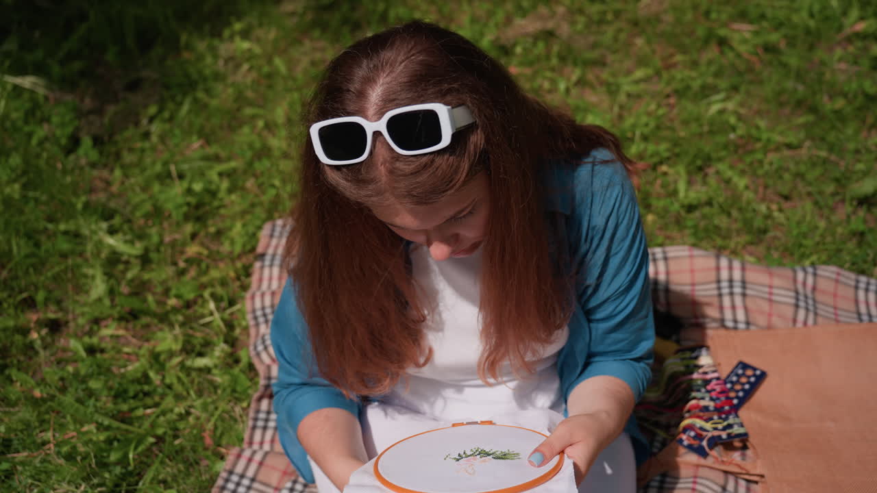Top down view of young girl sitting on blanket outdoors stitching fabric with focus and calm, sunlight illuminating her hair, surrounded by fresh green grass