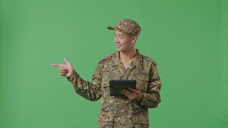 asian man soldier using a tablet and pointing to side while standing in the green screen background studio