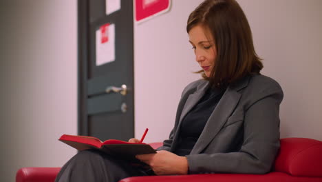 professional woman in gray suit writing notes in red notebook while sitting on red couch in office hallway, appearing focused and content, possibly preparing for meeting or reflecting ideas