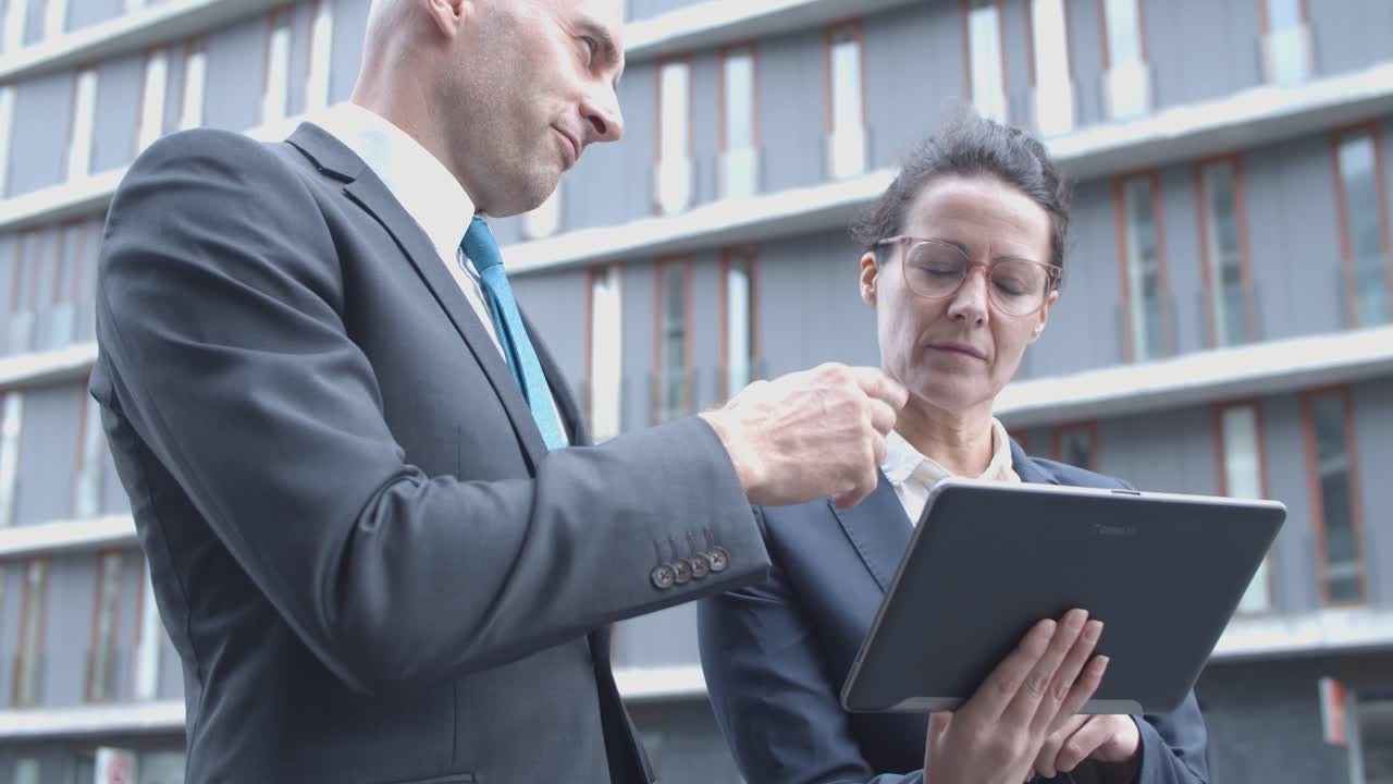 외부 사무실 건물 근처에 서 있는 동안 태블릿을 사용하여 프로젝트를 논의하는 두 비즈니스 파트너