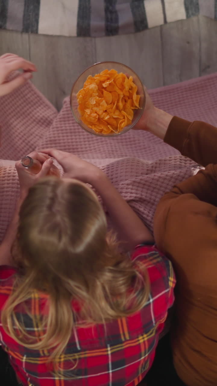 Long haired man treats women with chips watching TV series in apartment upper view slow motion. Friends chill out and enjoy good movie at home