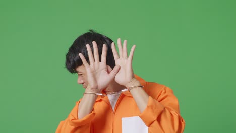 close up of asian male prisoner in handcuffs standing on the green screen background, showing palm sign the expression of not being wanted to be captured by camera