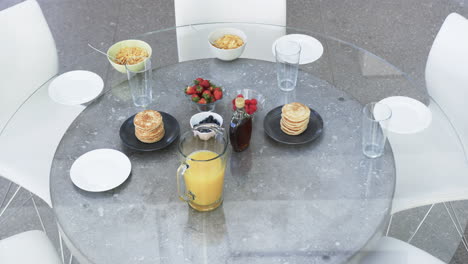 En-Una-Mesa-De-Cristal-Se-Sirve-Un-Desayuno-Con-Tortitas,-Frutas-Y-Zumo.