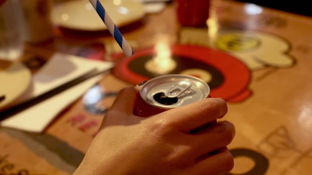 A hand inserts a blue-striped straw into a red soda can on a colorful table.