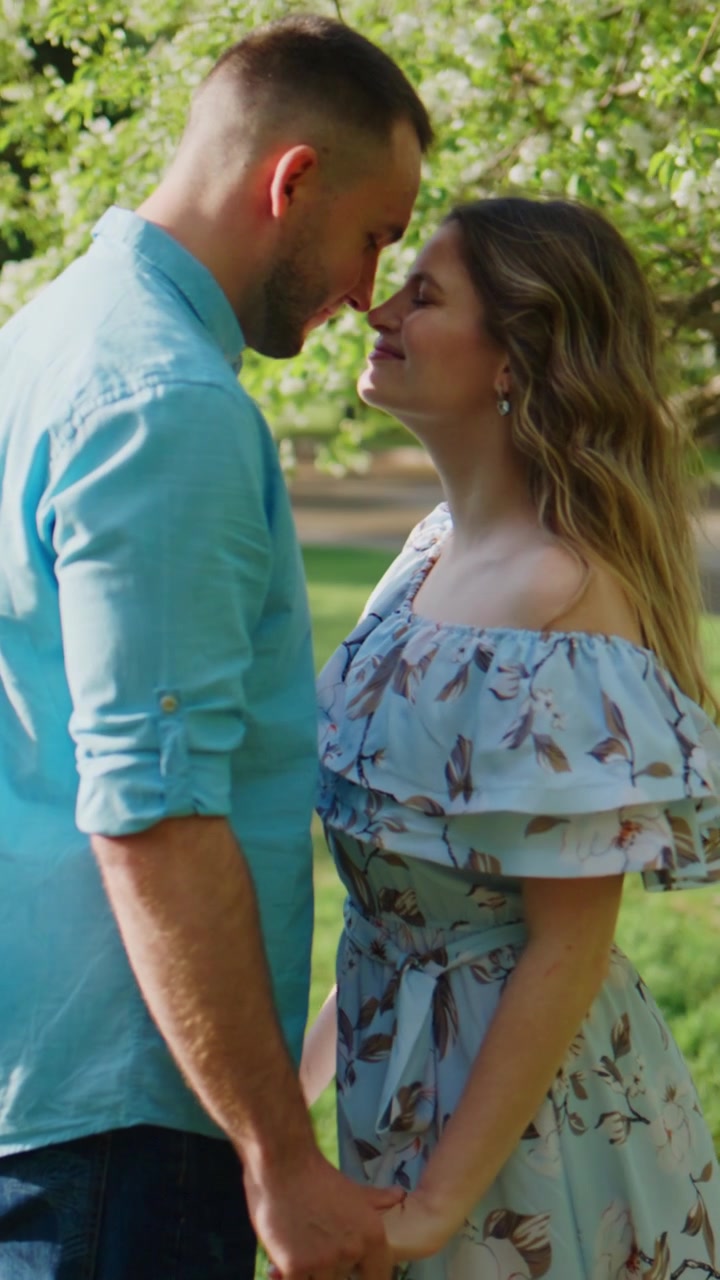 Casal de mãos dadas sob árvores floridas. O homem olha para a mulher com carinho. A mulher sorri, com os olhos fechados, desfrutando do momento. A luz suave do sol filtra-se pelas folhas verdes. Cena romântica perfeita para casamentos.