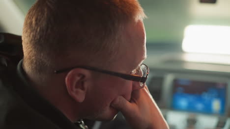 a close-up side profile of a man wearing glasses, seated inside a car, with his hand thoughtfully resting on his chin