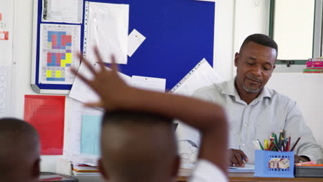 teacher smiles at kids from his desk in elementary class