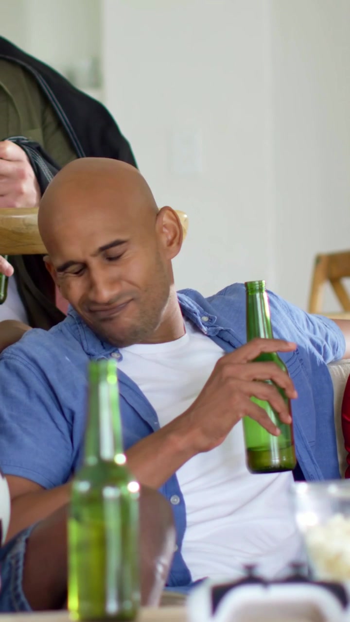 Video vertical: Hombre con camisa azul y cerveza girándose, inclinándose hacia un amigo que le da una palmada en el hombro en el sofá