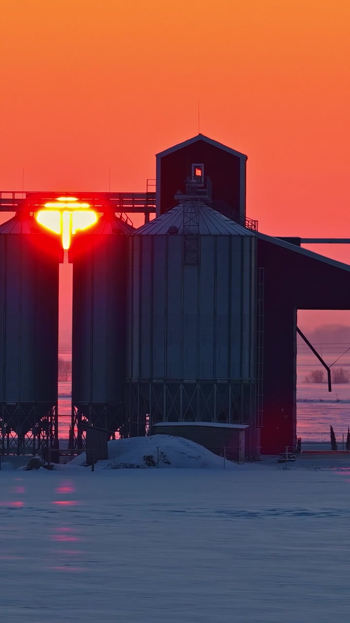 A cinematic drone flight of grain silos in a frozen rural landscape of Northern Europe at sunset, capturing ice and cold winter air, and dramatic golden light fading over industrial farm structures