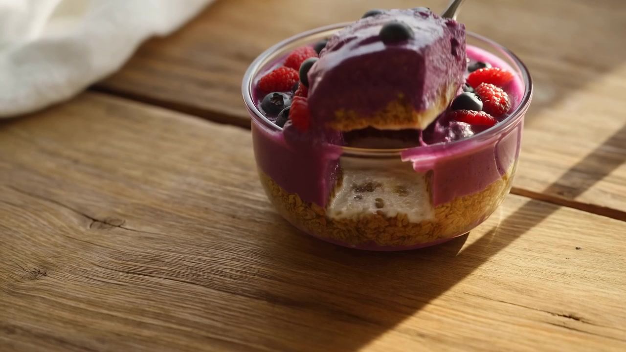 Close-up motion shows spoon removing scoop from layered berry yogurt parfait in glass bowl, leaving visible gap and creamy magenta mixture on spoon, with rustic wood surface, warm sunlight, fresh