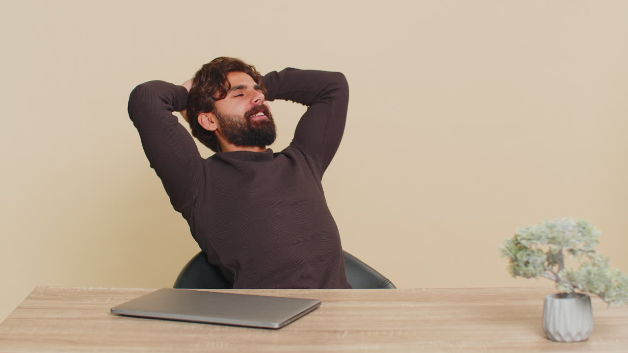 Happy young indian man closing laptop pc after finishing work at table against beige background