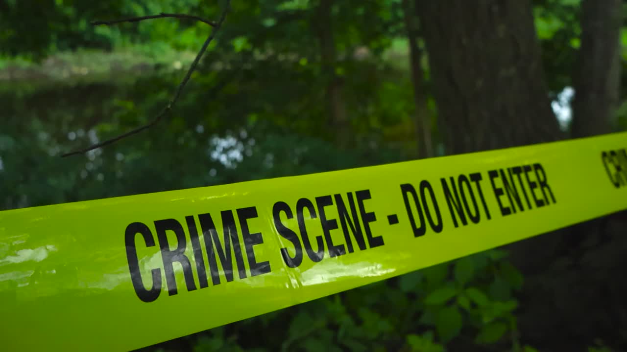 Yellow colored police line Crime Scene forensics investigation tape pulled in front of a day time thick forest area with a bokeh blurry lake visible in the background. Wind is moving the ribbon slowly