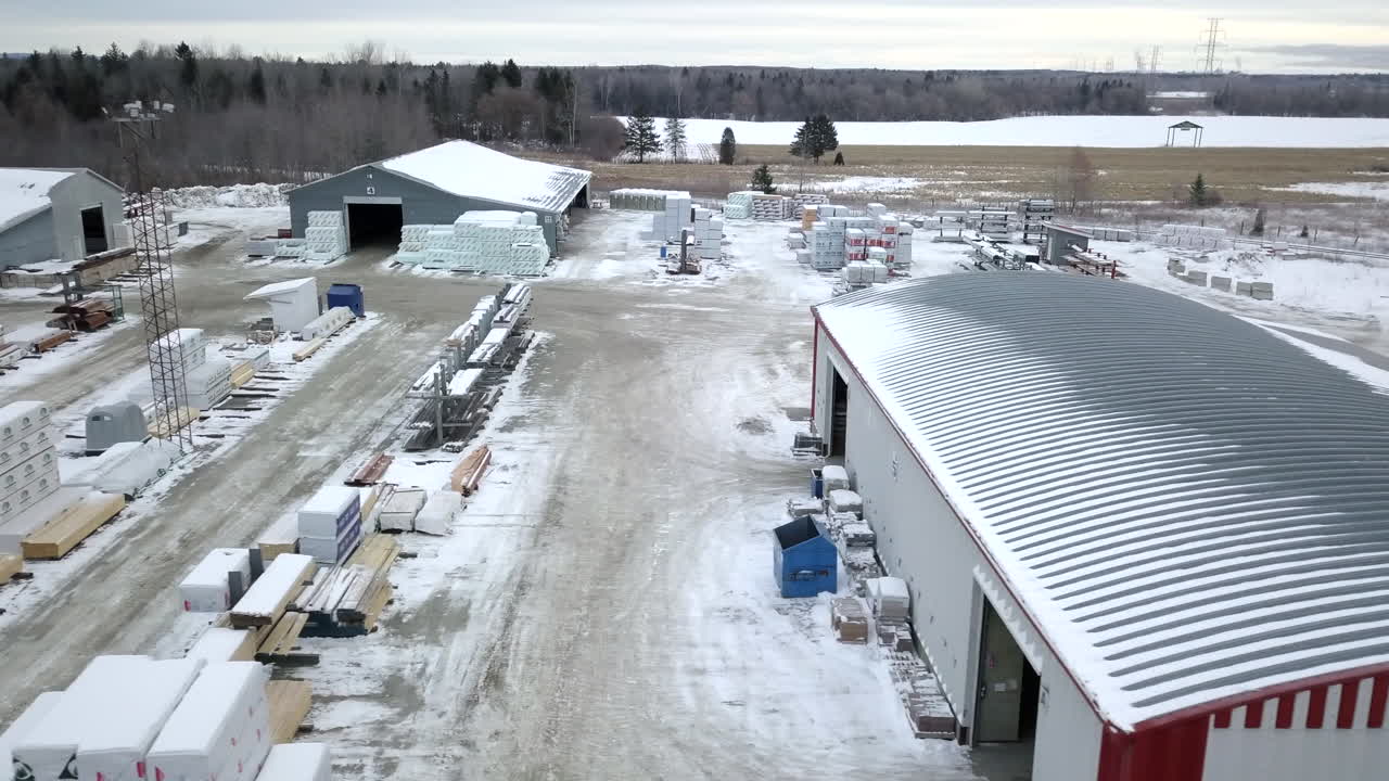 Piles Of Wood, Machineries, And Buildings In A Woodyard With Snow During Winter Season. aerial drone, sideways