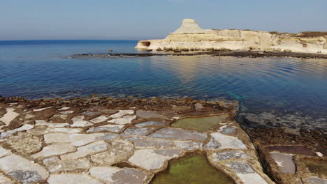 an aerial drone shot flying over the salt pans, rocks and crystal blue water of gozo island in malta