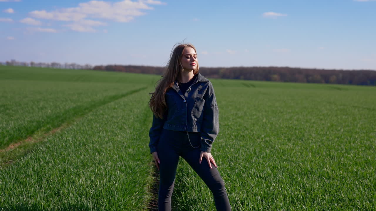 Attractive girl posing on camera outdoors. Beautiful young model in tight pants and cotton jacket with fluttering hair looking on camera on green field background.